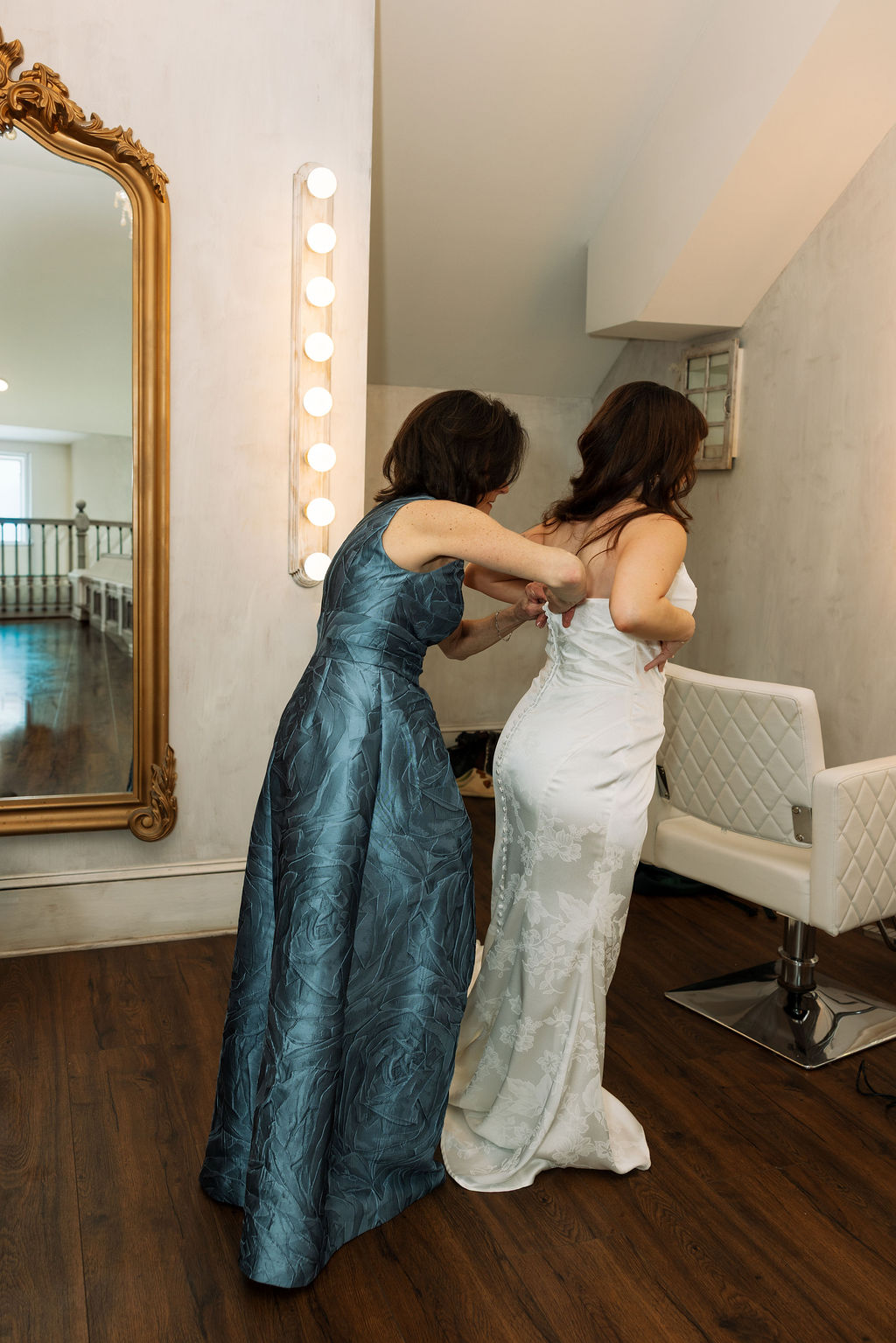 Mother helping button the back of the bride’s dress in a softly lit getting ready space