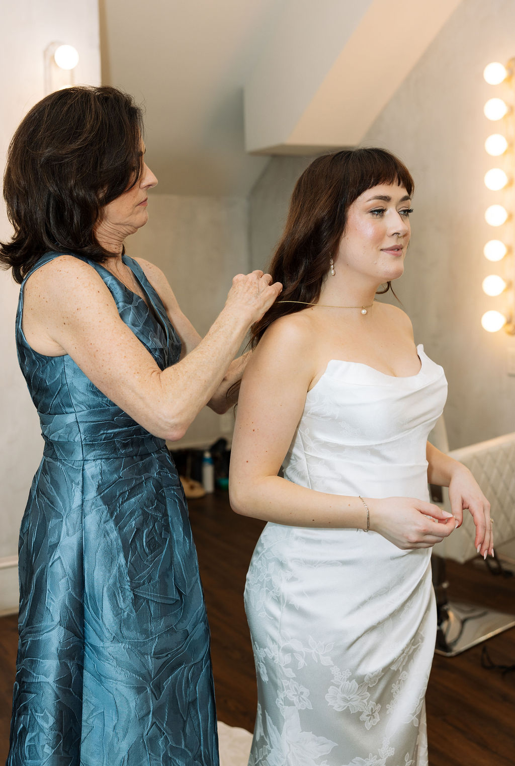 Mother fastening the bride’s necklace in a softly lit getting ready room