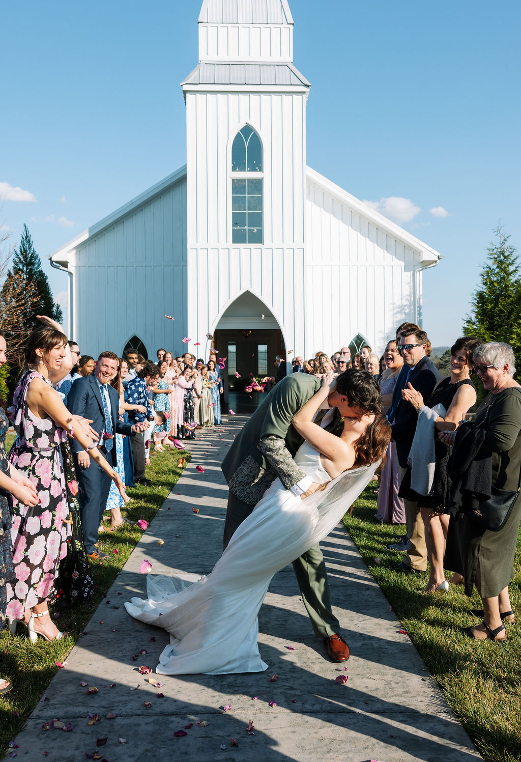 Couple sharing a dip kiss outside the white chapel surrounded by cheering guests at Howe Farms wedding venue