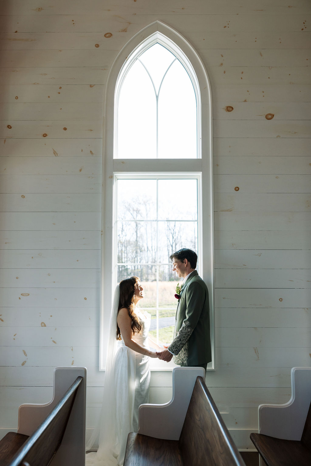 Bride and groom holding hands in front of a tall arched window inside the chapel at Howe Farms wedding venue