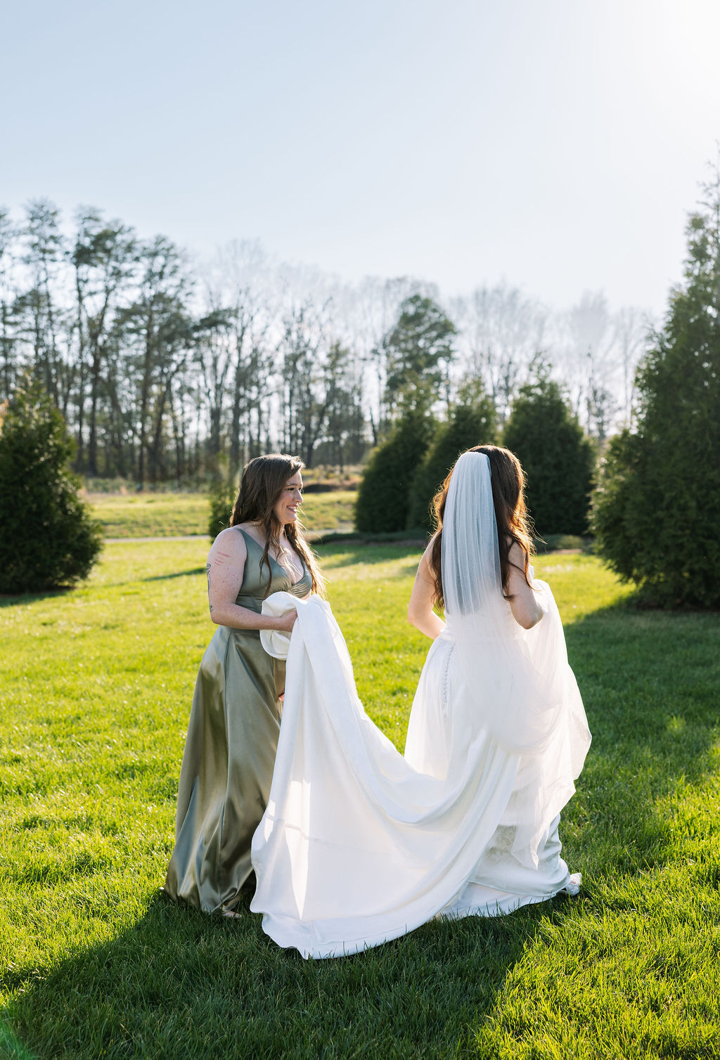 Bride and bridesmaid walking through a sunlit field, carrying the train of her dress at Howe Farms wedding venue