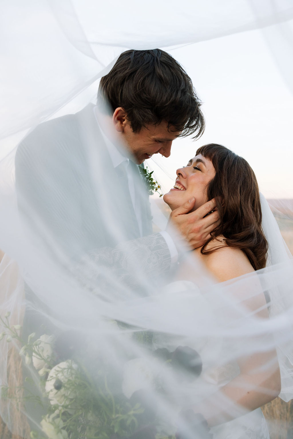 Couple wrapped in the bride’s veil, smiling and leaning into each other outdoors