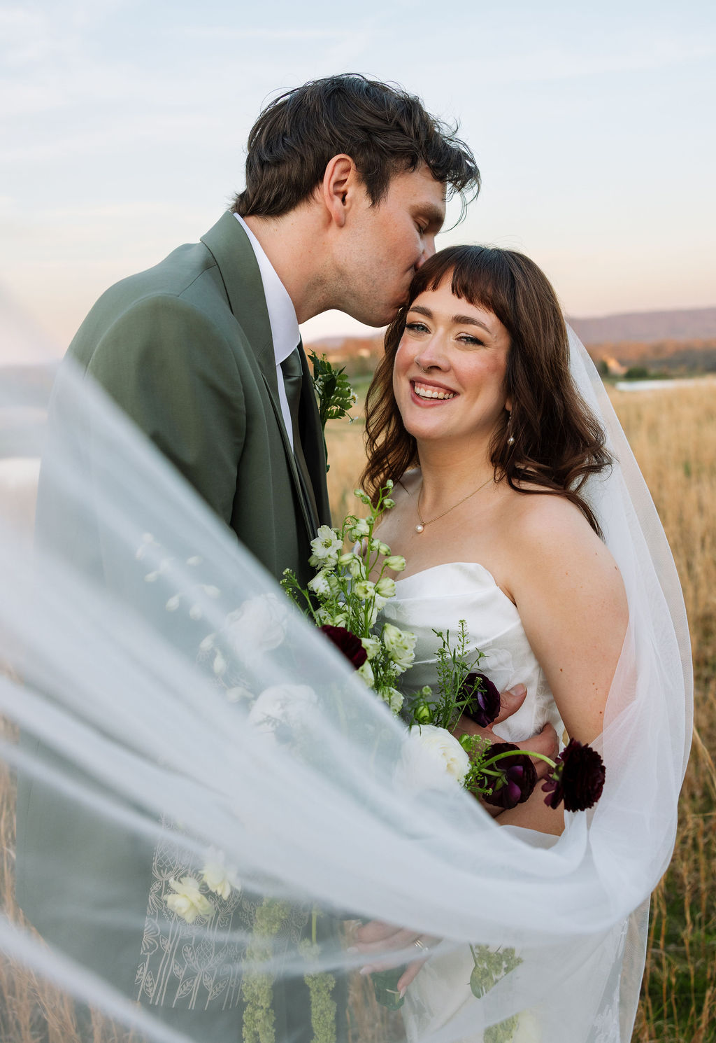 Groom kissing the bride’s forehead as her veil moves softly in the breeze