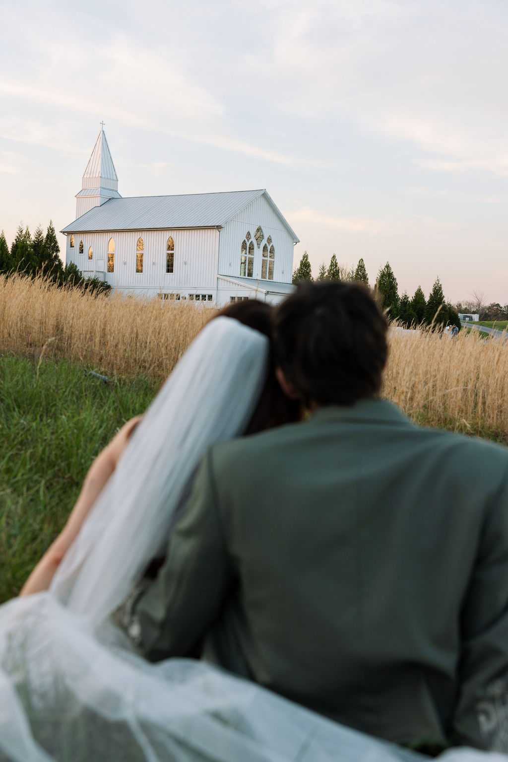 Newlyweds sitting together in a field looking toward the white chapel at Howe Farms wedding venue