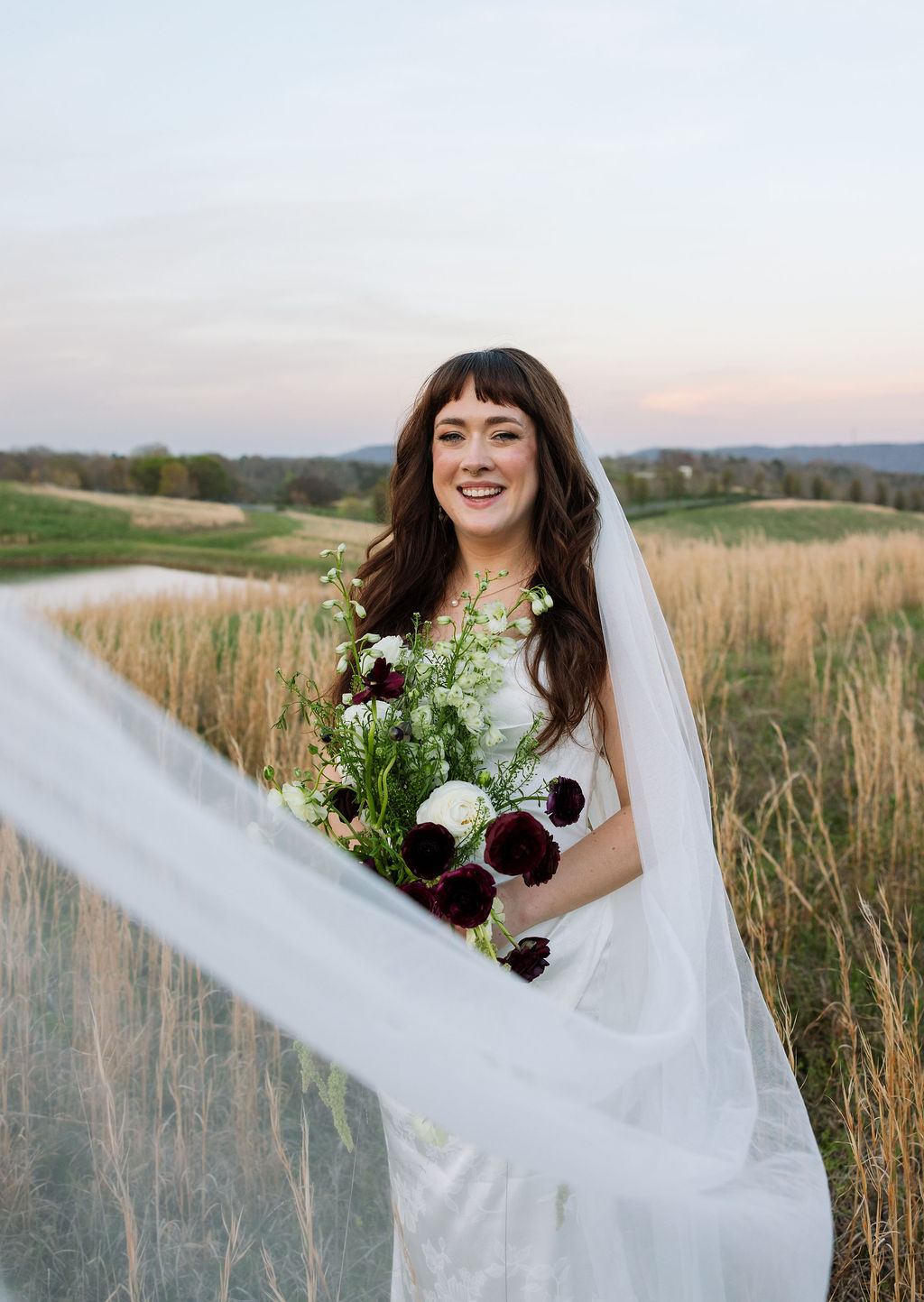 Bride holding her bouquet in a field with her veil moving softly in the wind at Howe Farms wedding venue