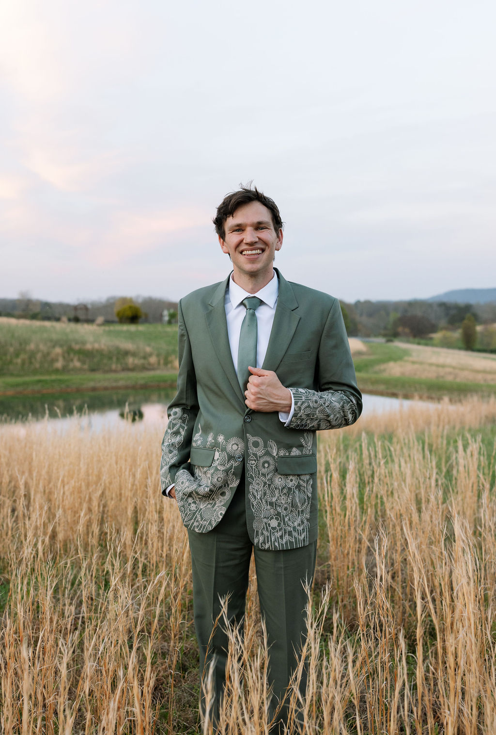 Groom standing in tall grass, smiling in a green suit with rolling hills behind him