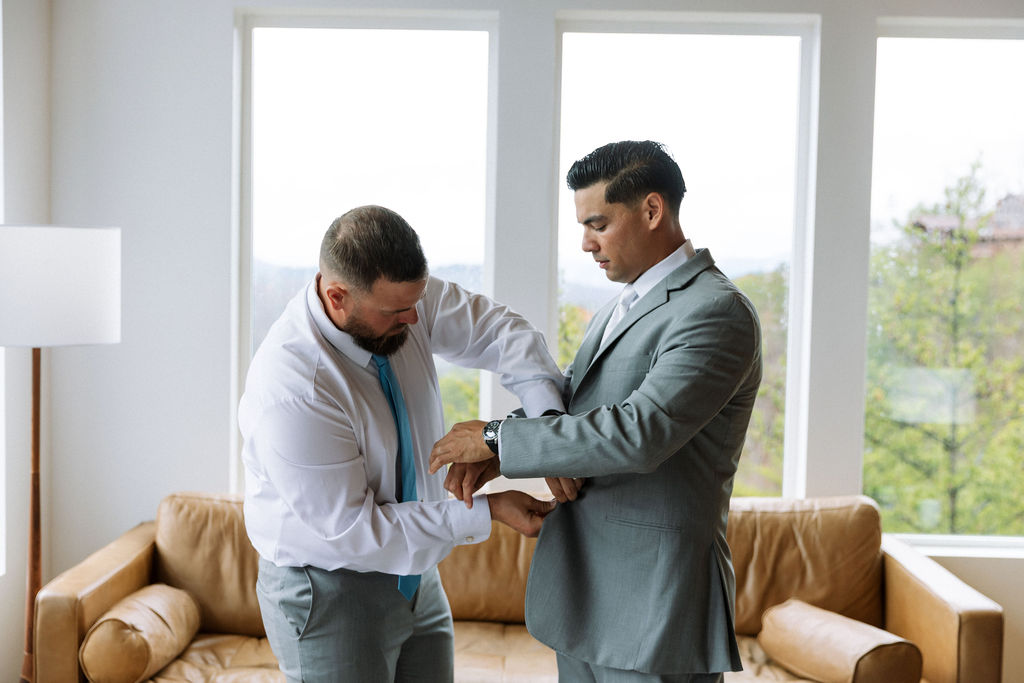 Groom getting ready with help adjusting his cuff and jacket in a bright room with mountain views
