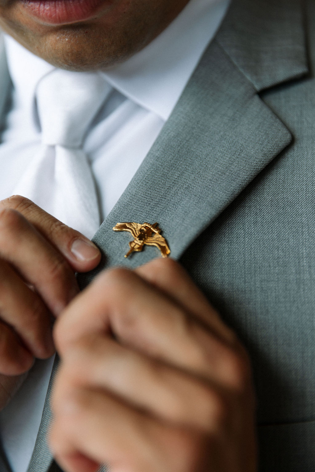 Close-up of groom adjusting a gold bird lapel pin on a light gray suit jacket

