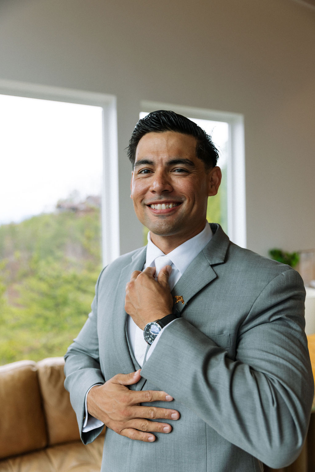 Groom smiling while adjusting his tie in a light-filled room with mountain views
