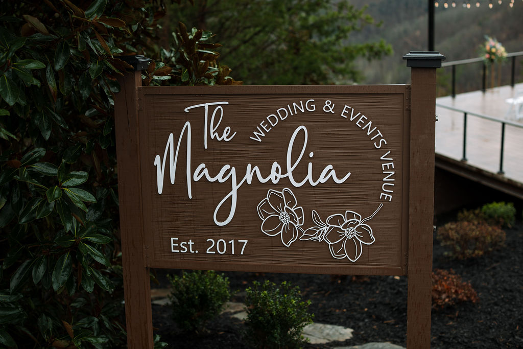 Wooden welcome sign for The Magnolia Wedding Venue surrounded by greenery and overlooking the ceremony space
