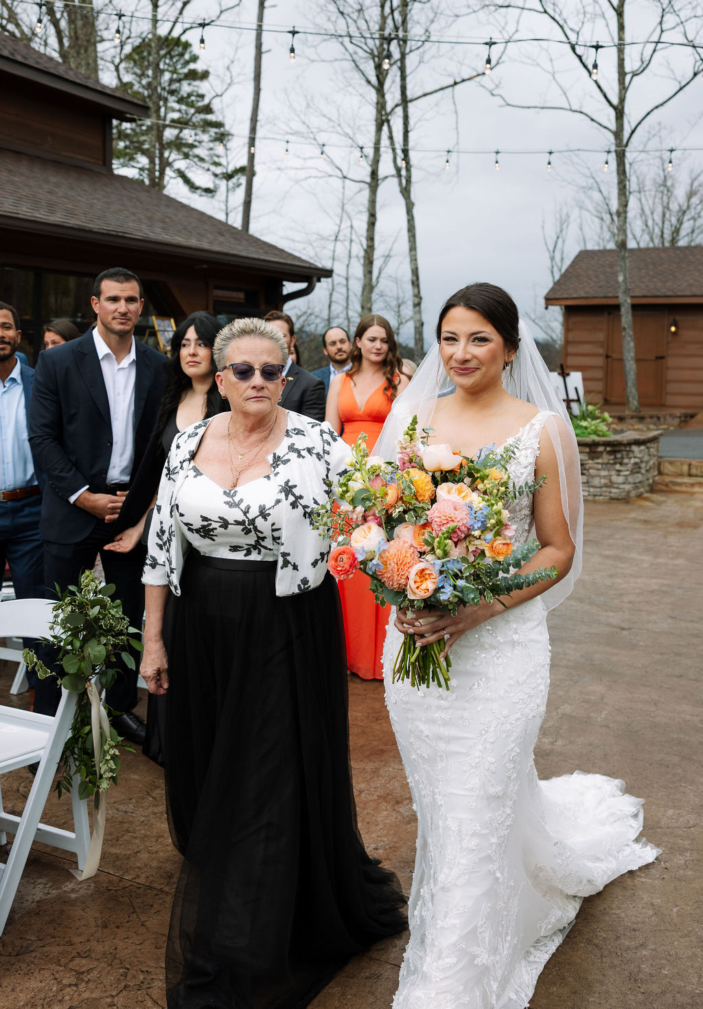 Bride walking down the aisle with a loved one, holding a colorful bouquet during an outdoor ceremony
