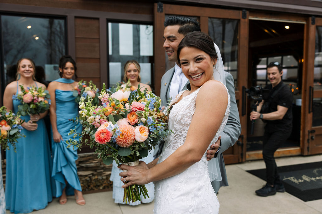 Bride laughing and holding bouquet surrounded by bridesmaids outside the reception space
