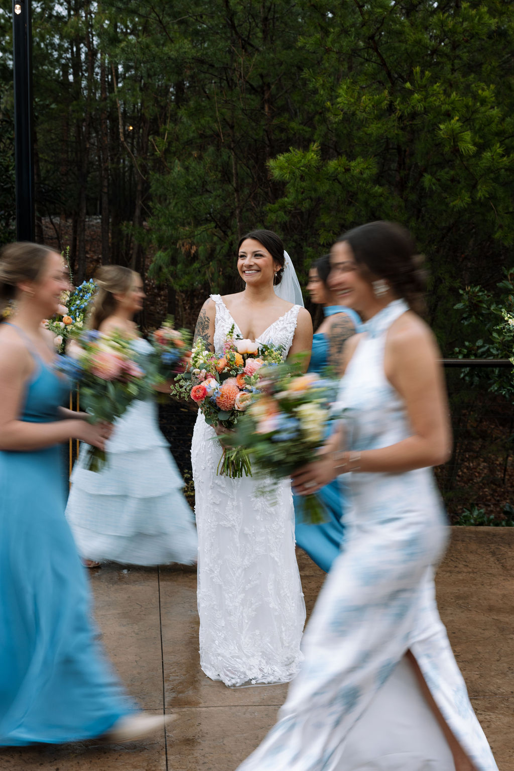 Bride standing still with bouquet while bridesmaids move around her in soft motion outdoors
