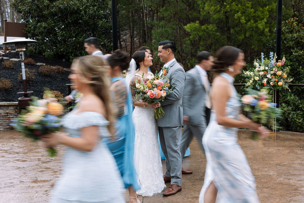 Bride and groom pause together as wedding party moves around them in soft motion blur during an outdoor celebration
