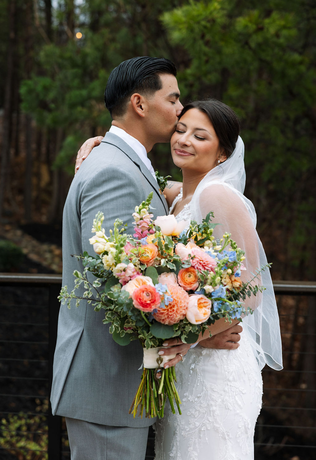 Bride and groom embrace outdoors at The Magnolia Wedding Venue, surrounded by pine trees, holding a vibrant bouquet
