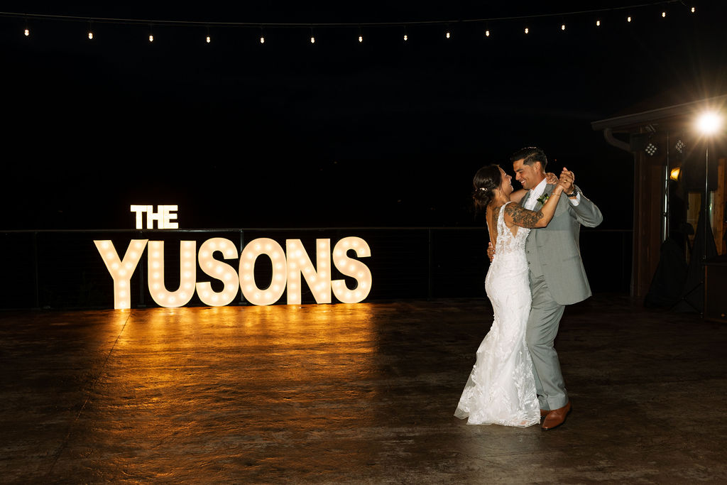 Bride and groom dancing under string lights with custom wedding signage at night
