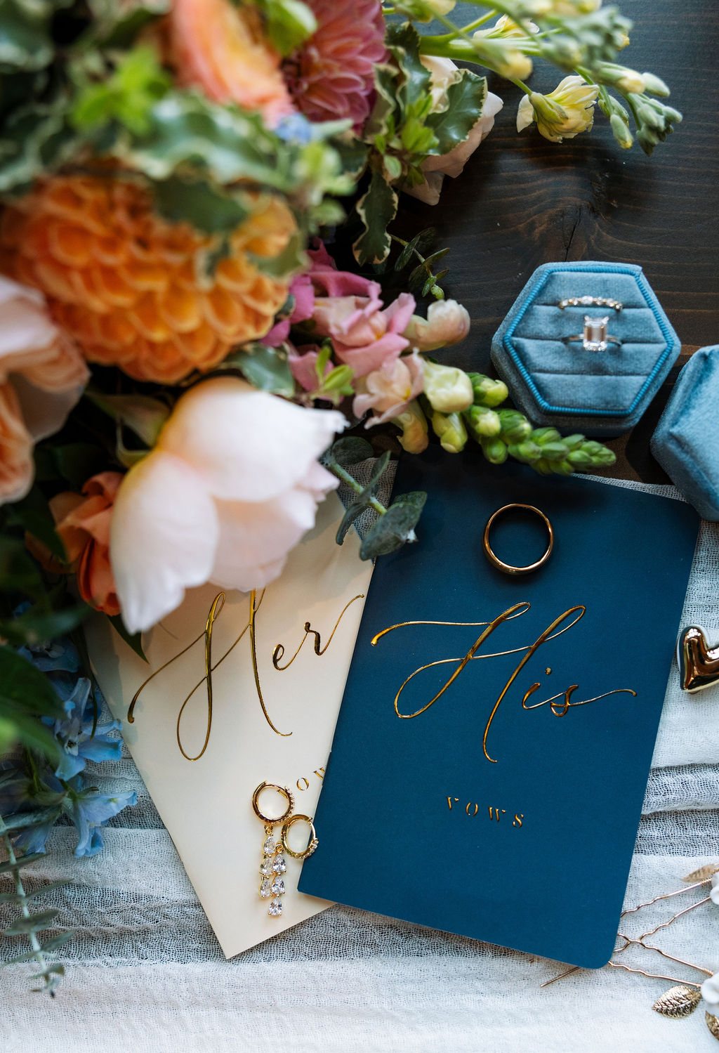 Wedding invitation suite with blue velvet ring box, gold jewelry, and colorful florals arranged on soft fabric
