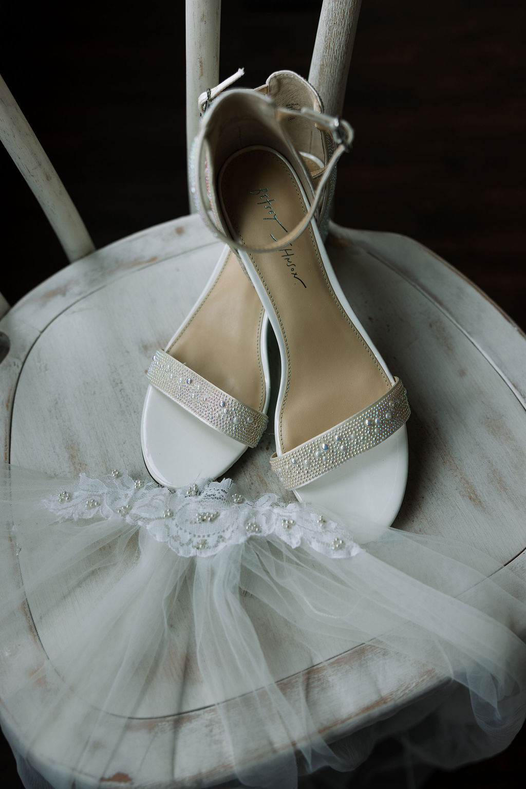 Bridal shoes with pearl detailing and lace garter placed on a white chair in soft window light
