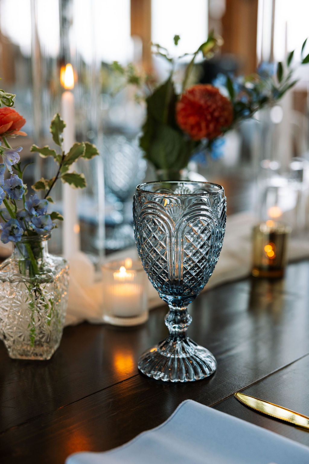 Close-up of a textured blue goblet surrounded by candles and delicate floral arrangements on a dark wood reception table