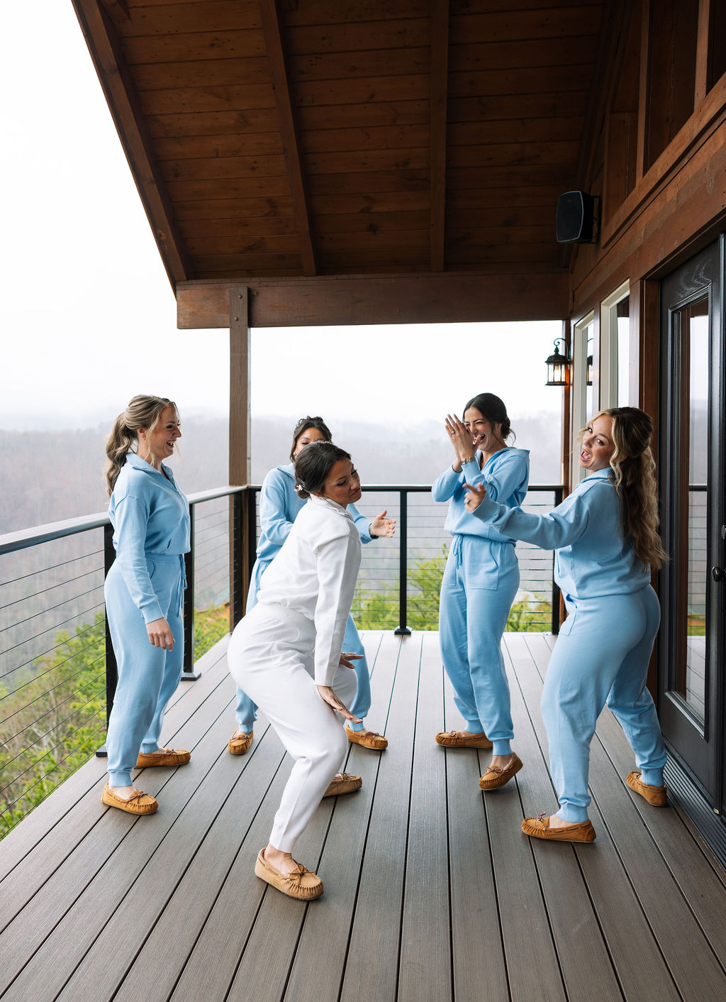 Bride and bridesmaids in matching blue outfits dancing together on a covered deck at The Magnolia Wedding Venue
