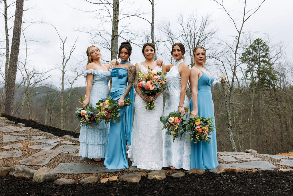 Bridal party standing together on a mountain overlook with blue dresses and colorful bouquets
