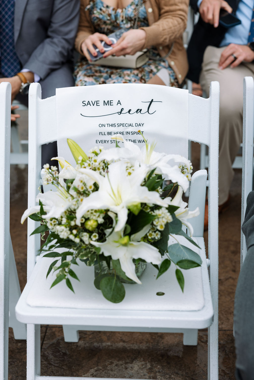 Reserved ceremony chair with white florals and a sign reading “save me a seat” during an outdoor wedding
