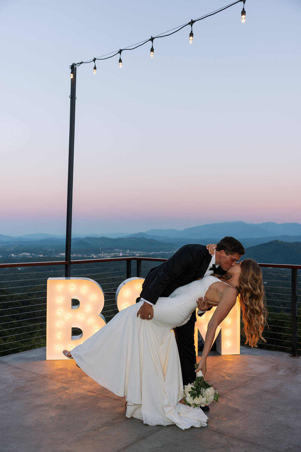 Bride and groom dipping kiss under string lights with mountain views during Knoxville wedding venue reception