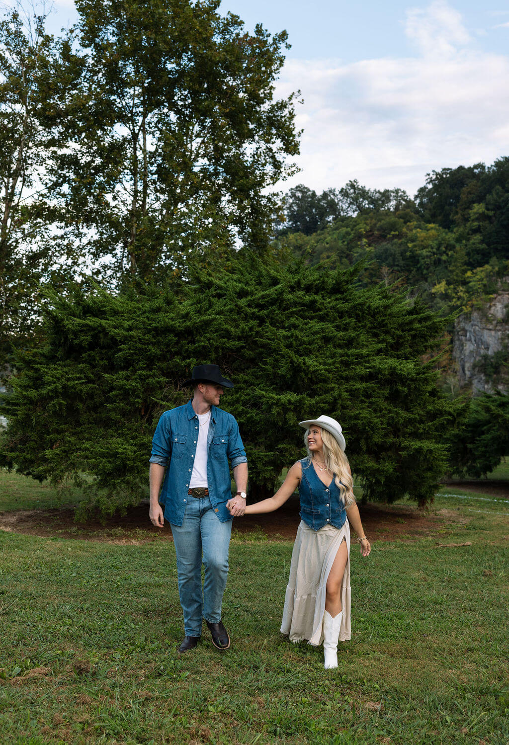 Couple walking hand in hand through a grassy field during a Knoxville, Tennessee engagement session at sunset.