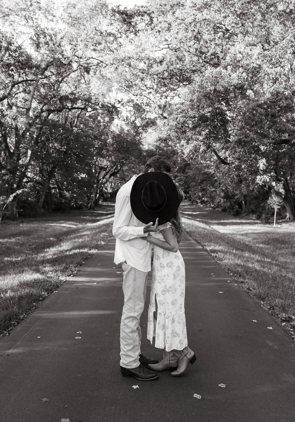Black and white engagement portrait of couple embracing on a tree lined road at Maple Grove Estate