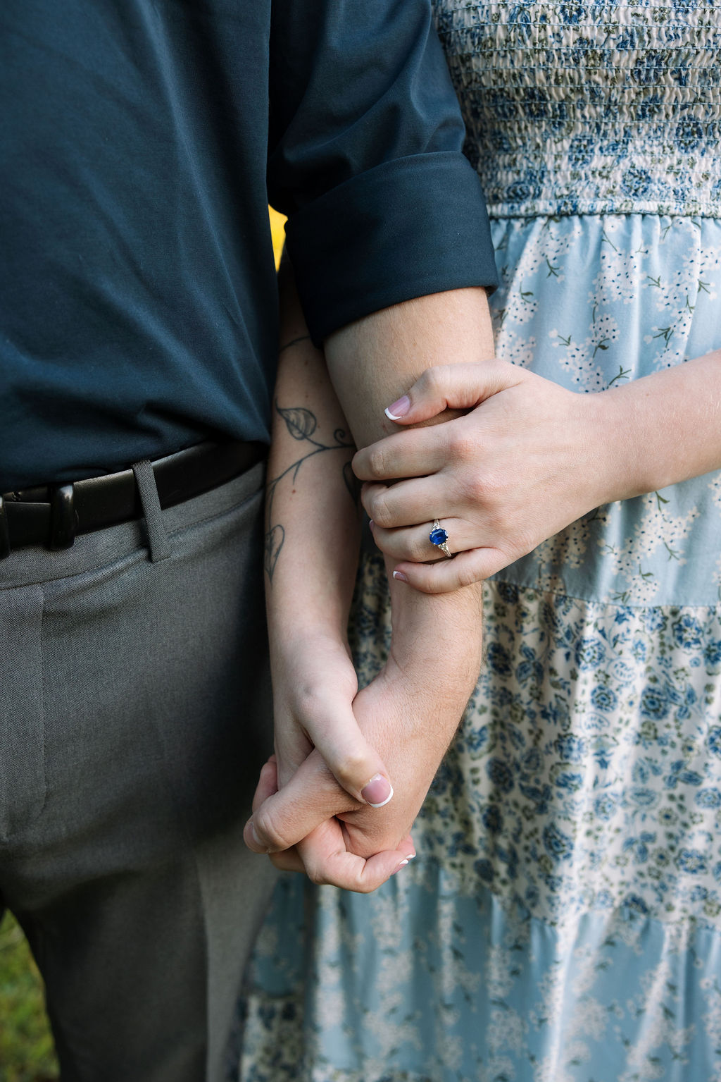 Close-up detail of an engaged couple holding hands, showing the woman's sapphire engagement ring against her floral blue dress at Knoxville Botanical Garden.