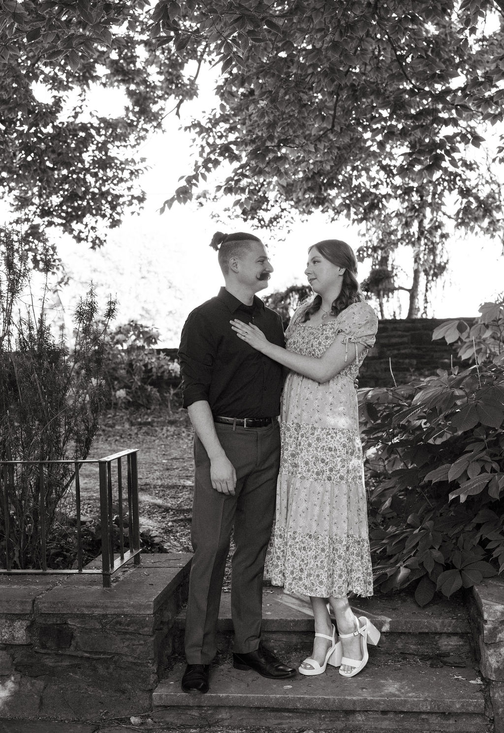 Black and white photo of an engaged couple laughing together on a stone staircase at Knoxville Botanical Garden, the woman in a floral dress and heels looking up at her partner.
