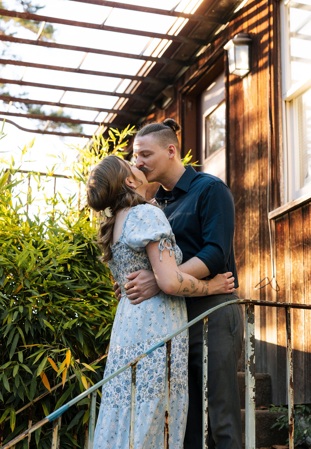 Couple kissing outside the Stone Greenhouse at Knoxville Botanical Garden, the woman in a blue floral dress with her arms around her partner beside lush bamboo and warm evening light.