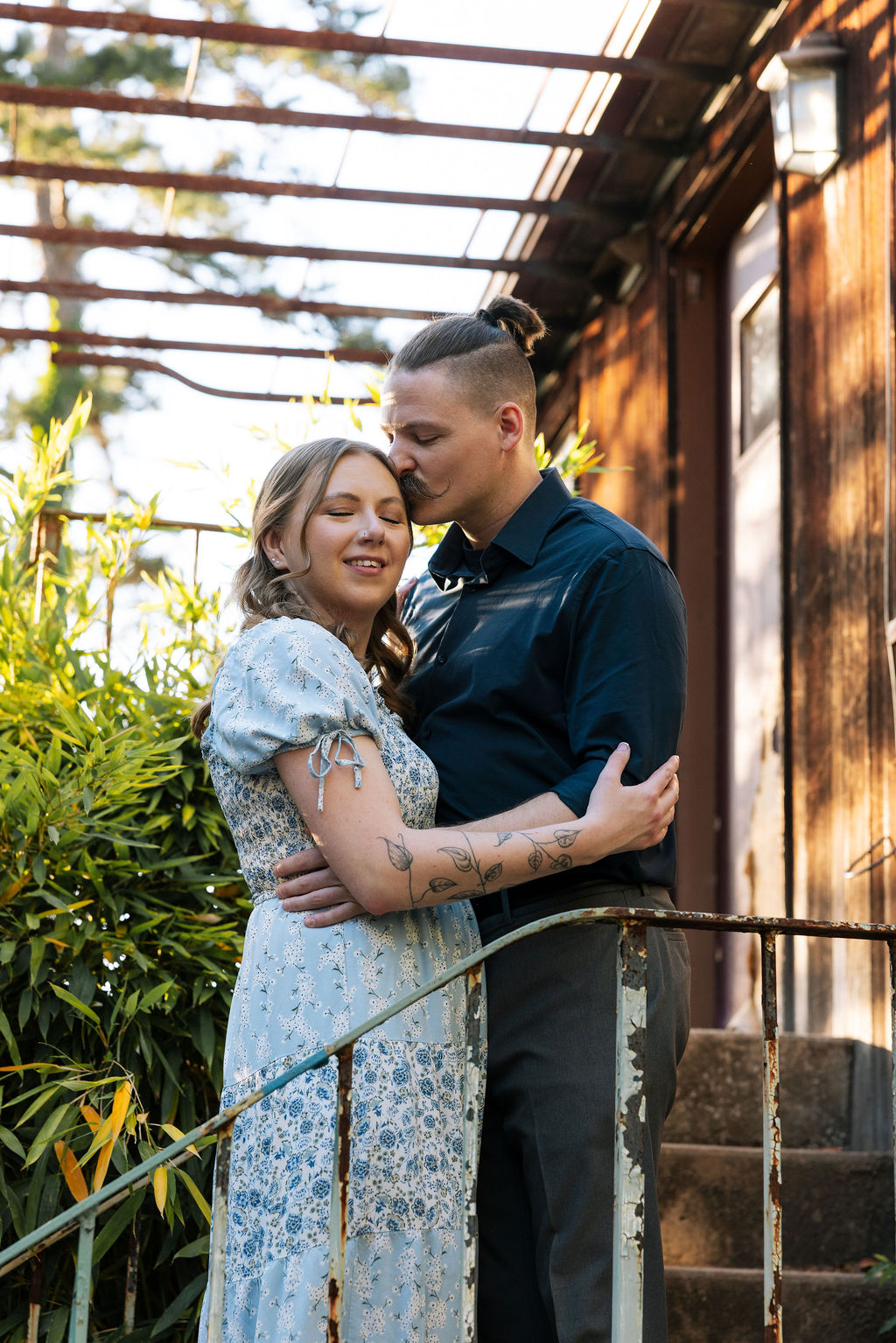 Couple standing close together outside the historic Stone Greenhouse at Knoxville Botanical Garden, the woman in a floral blue dress smiling as her partner leans in during their engagement session.