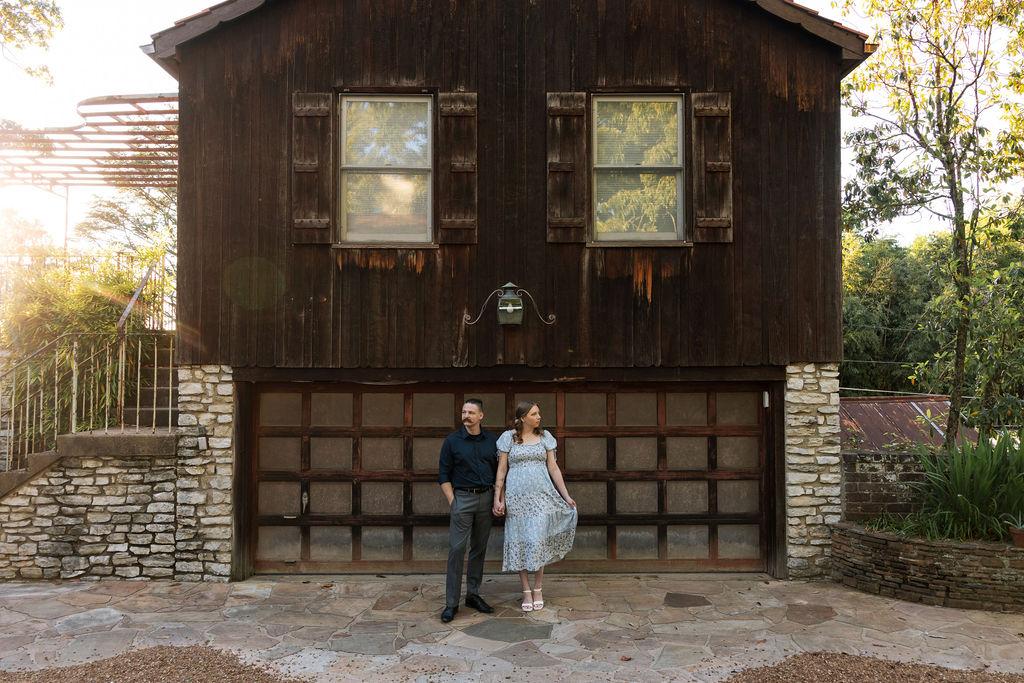 Wide shot of an engaged couple standing in front of the iconic dark wood barn at Knoxville Botanical Garden at sunset, the rustic structure and stone foundation glowing in the golden light with the pergola and lush trees visible on either side.