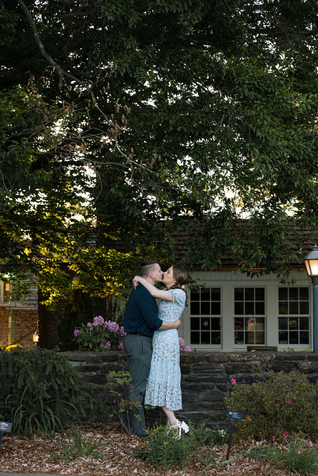 Couple kissing in front of a stone cottage surrounded by garden beds and mature trees at Knoxville Botanical Garden during a warm summer engagement session.