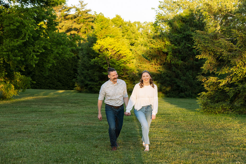 Couple walking through a wide open lawn at Knoxville Botanical Garden during golden hour, hand in hand and smiling, surrounded by tall glowing green trees lit by the setting sun.