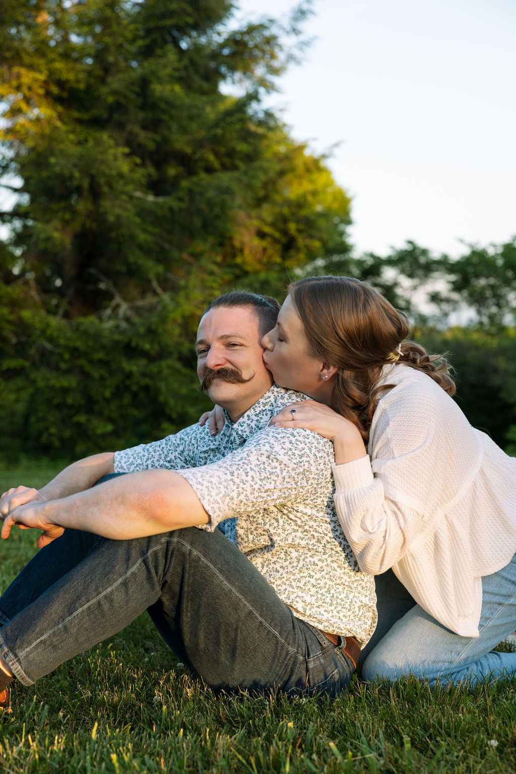Couple sitting in the grass at Knoxville Botanical Garden, the man leaning in close and whispering to his partner as she smiles, both relaxed and laughing in soft golden light.