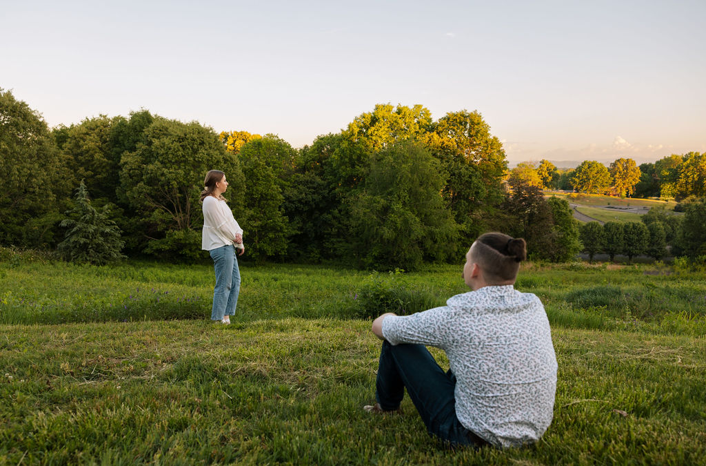 Wide editorial shot of an engaged couple at Knoxville Botanical Garden during golden hour, the man sitting in the grass in the foreground as his partner stands in the distance looking out over a sweeping green meadow and treeline.