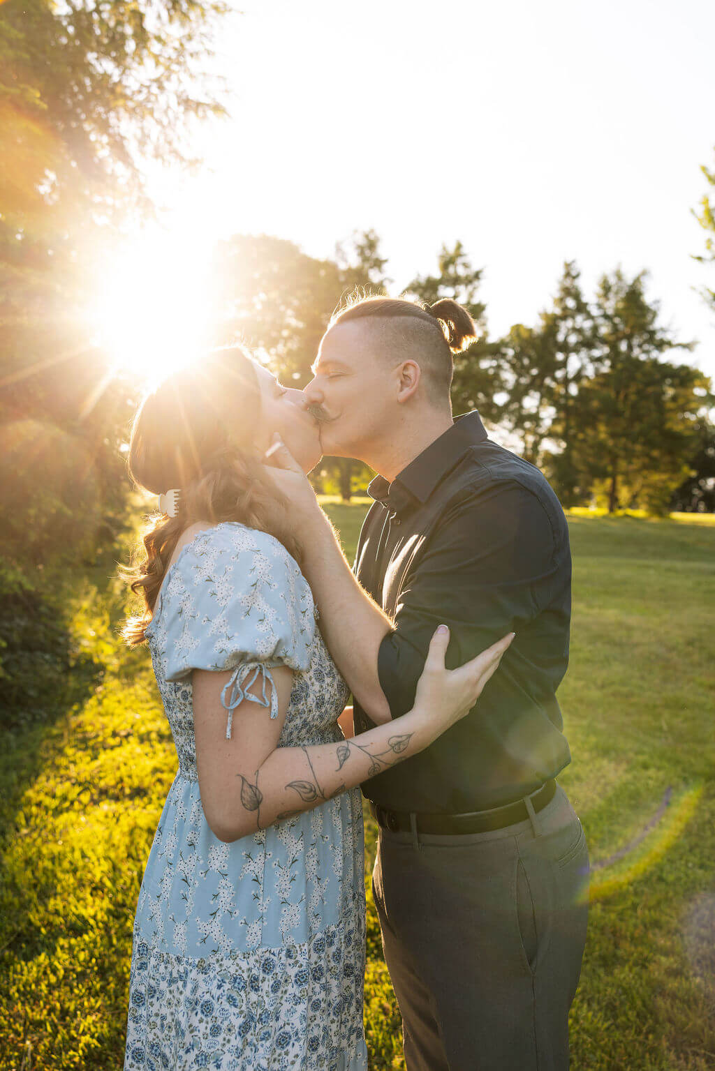 Couple kissing during golden hour at Knoxville Botanical Garden, the woman in a blue floral dress with her hand on her partner's chest, warm sun flare surrounding them in the open field.