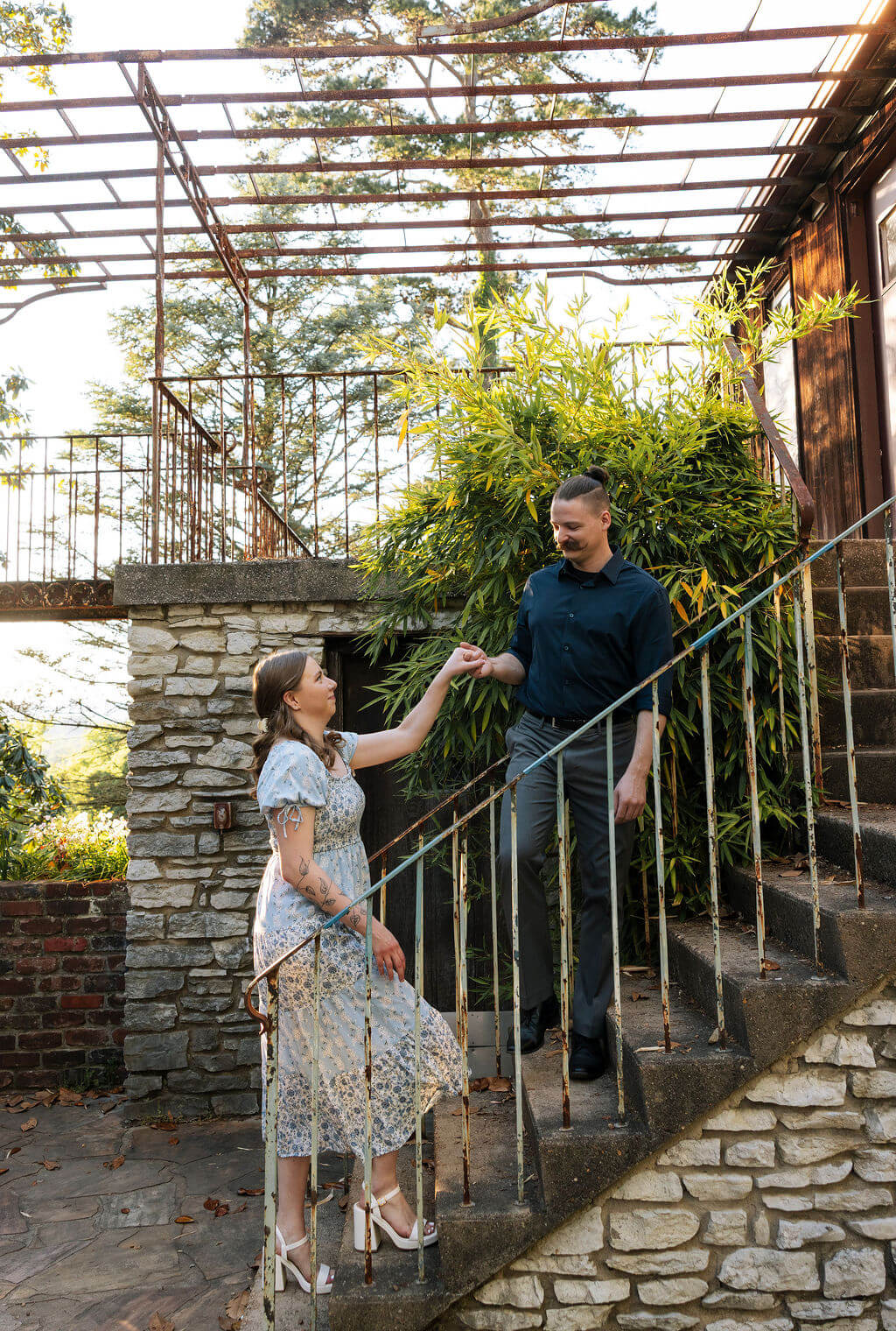 Engaged couple holding hands on a stone staircase covered in climbing vines at Knoxville Botanical Garden, the woman in a floral midi dress looking up at her partner.