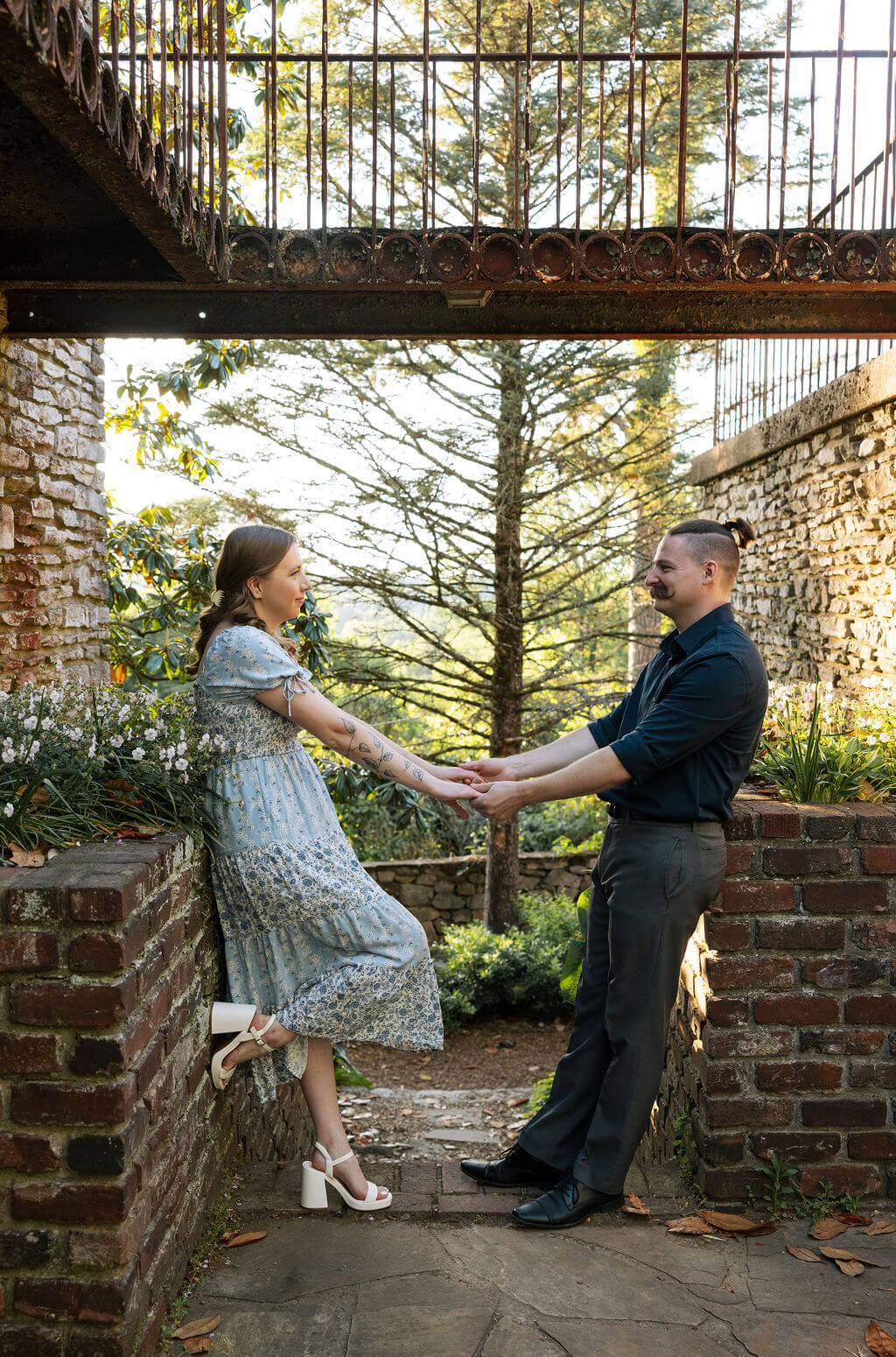 Engaged couple holding hands near a weathered iron gate and brick wall at Knoxville Botanical Garden, the woman in a flowy blue dress standing on a step as they face each other in warm evening light.