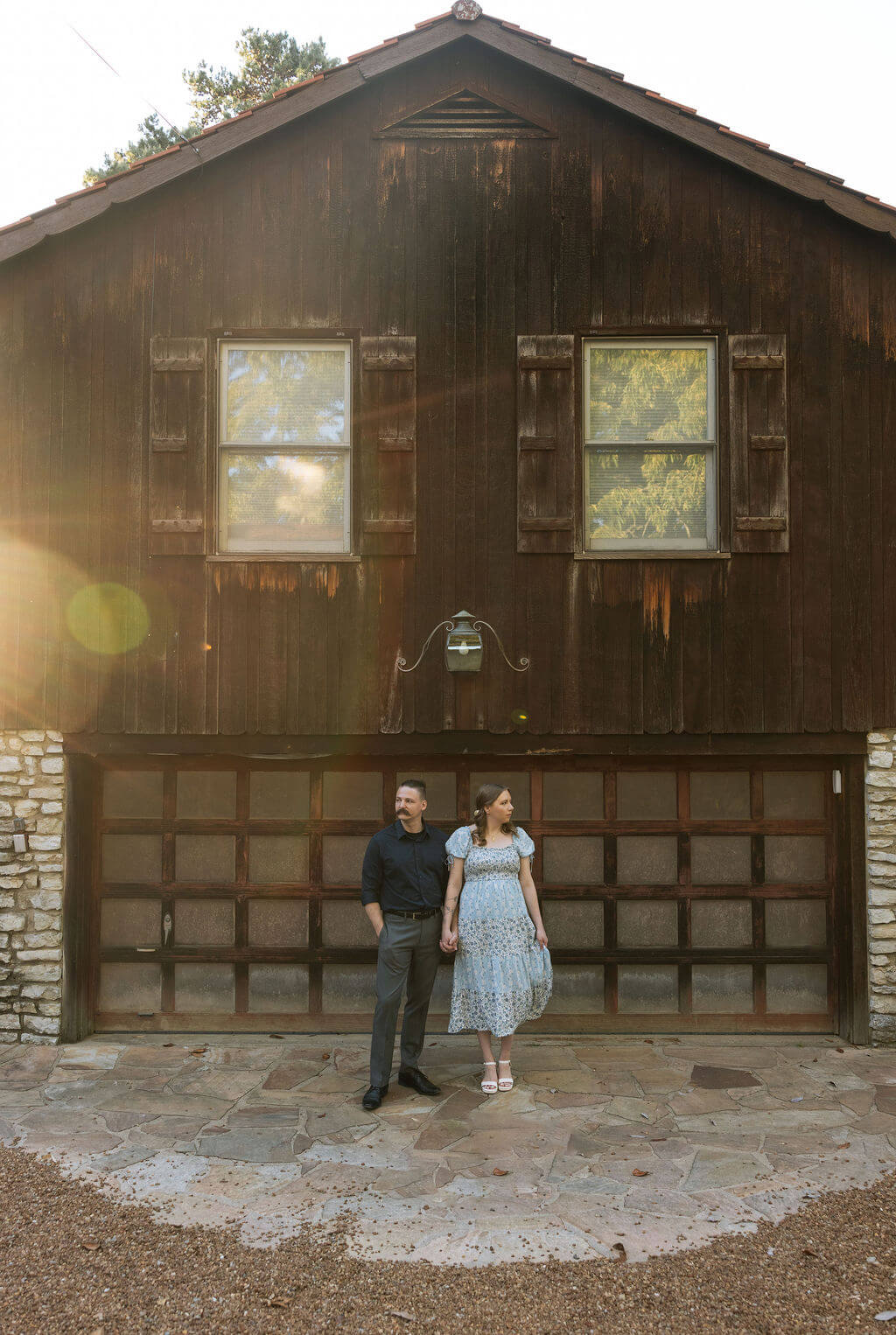 Couple standing in front of a rustic dark wood barn at Knoxville Botanical Garden during golden hour, stone details and warm lens flare framing the scene behind them.