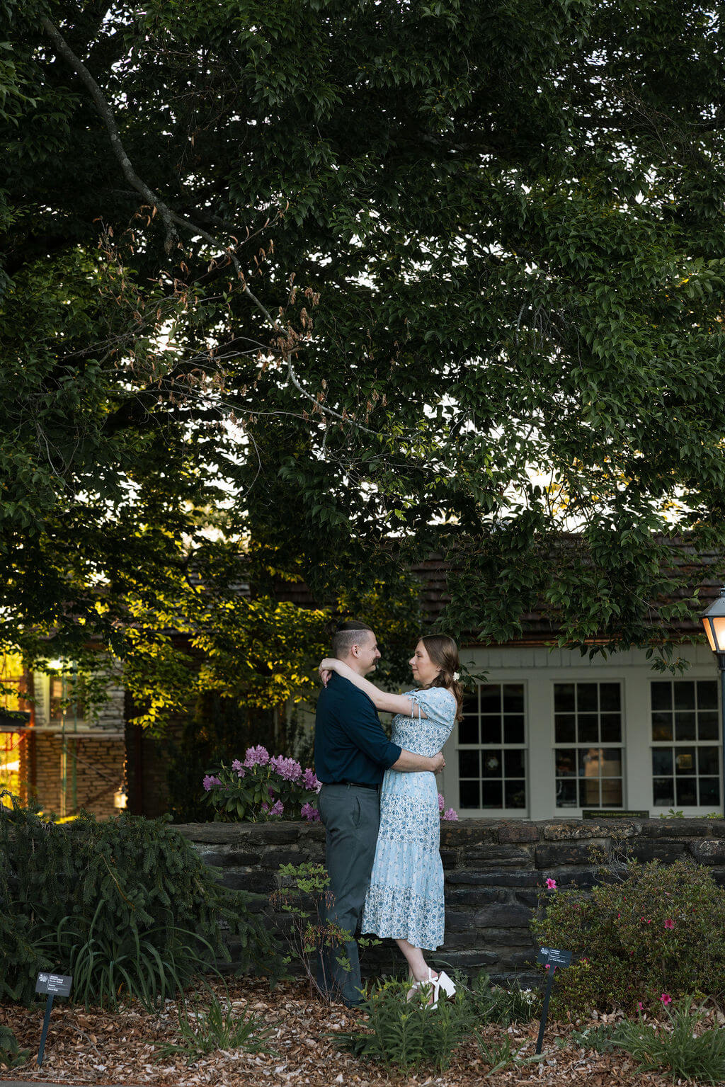 Couple embracing near a stone cottage at Knoxville Botanical Garden at dusk, surrounded by lush garden beds and large mature trees with warm light glowing from the windows behind them.