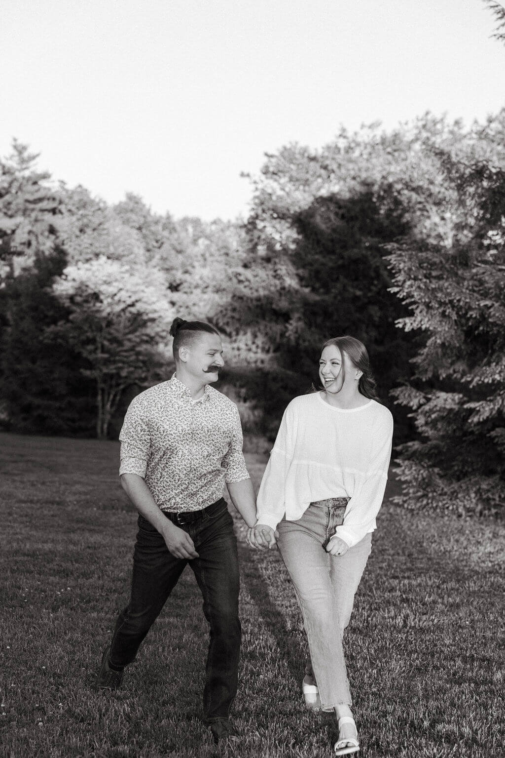 Black and white photo of an engaged couple laughing and being playful while walking through an open lawn at Knoxville Botanical Garden, the woman in a white top and jeans mid-step.