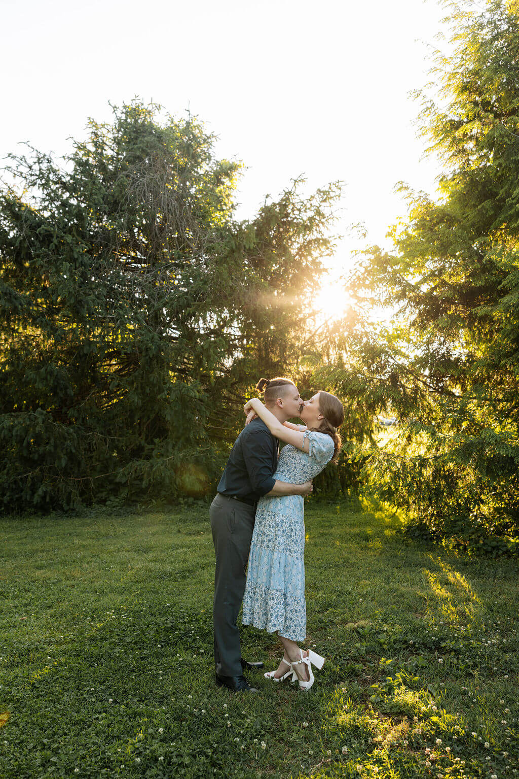 Couple sharing a kiss while standing in an open grassy area at Knoxville Botanical Garden, backlit by warm sunset light filtering through the trees.