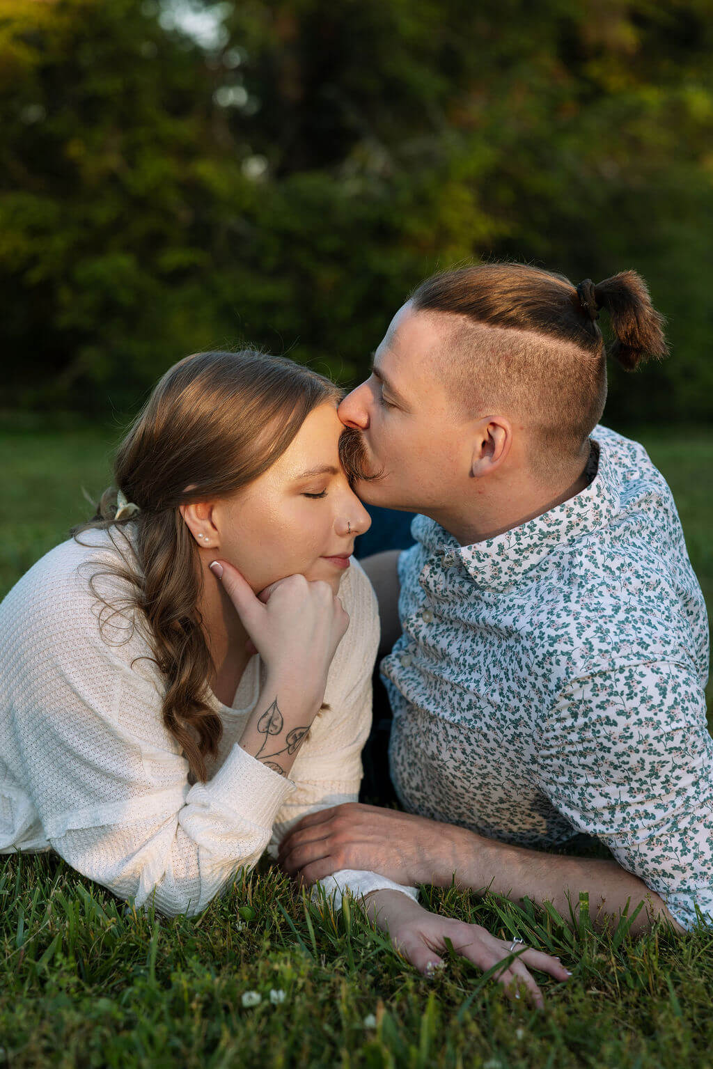Couple lying in the grass together at Knoxville Botanical Garden, the man leaning in to kiss the woman on the cheek during their engagement session in golden evening light.