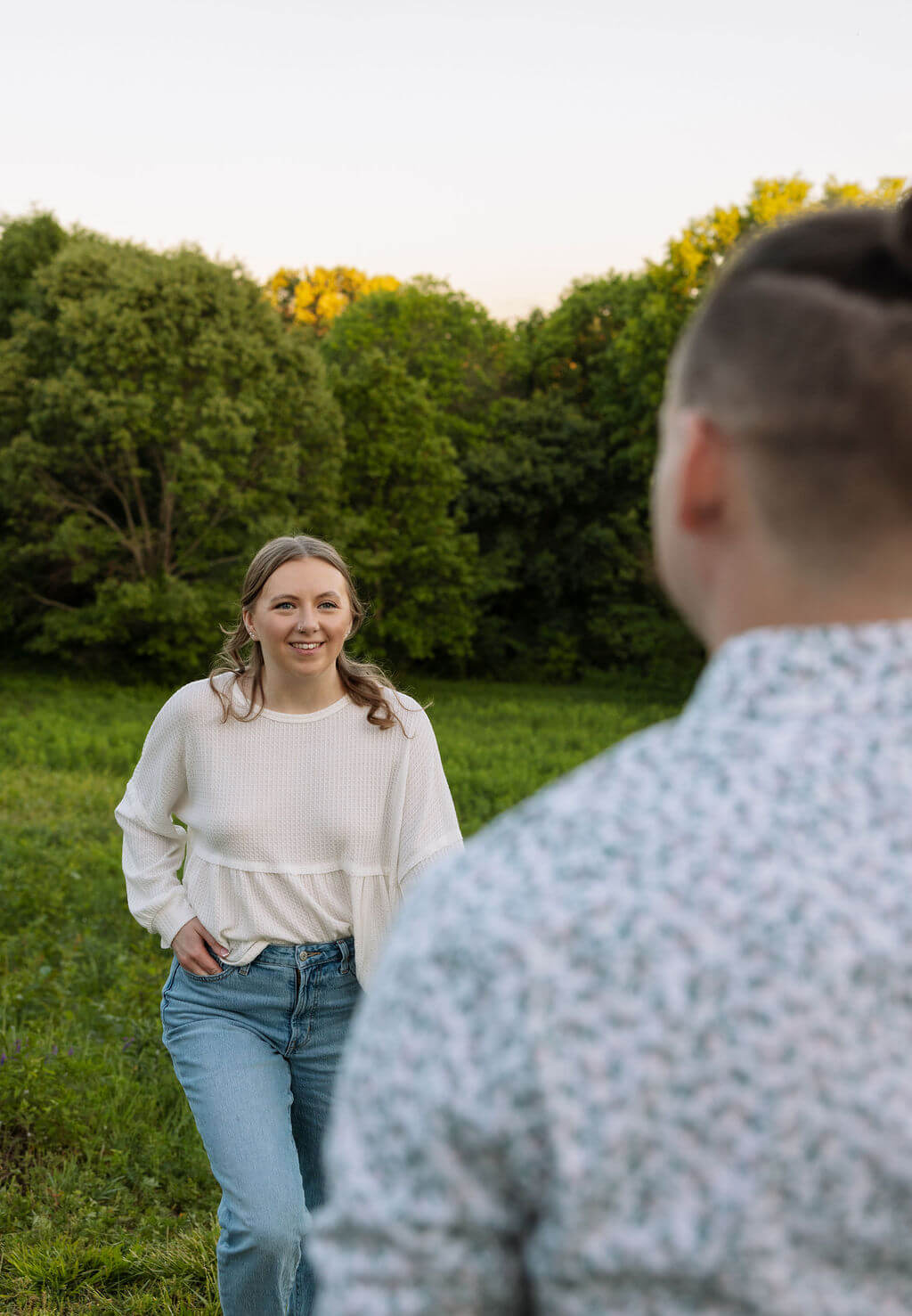 Man standing in a meadow at Knoxville Botanical Garden looking back at his partner walking toward him through golden hour light, surrounded by lush green trees.