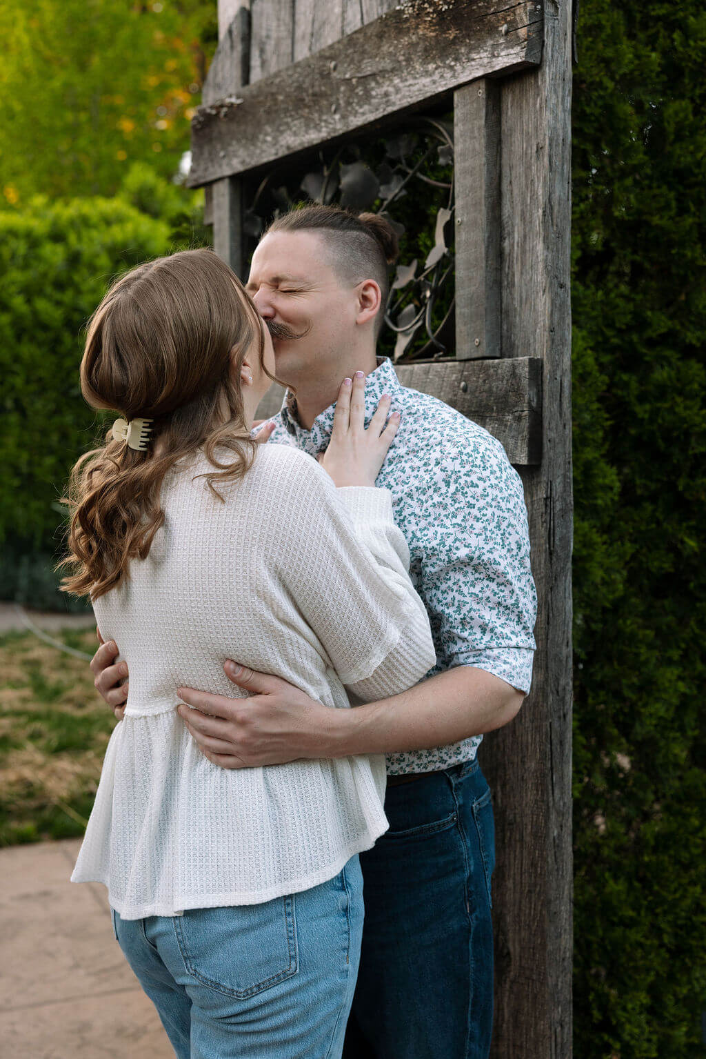 Couple sharing a tender kiss beside a rustic wooden structure at Knoxville Botanical Garden, the woman in a cream sweater with her hand on her partner's chest in soft evening light.