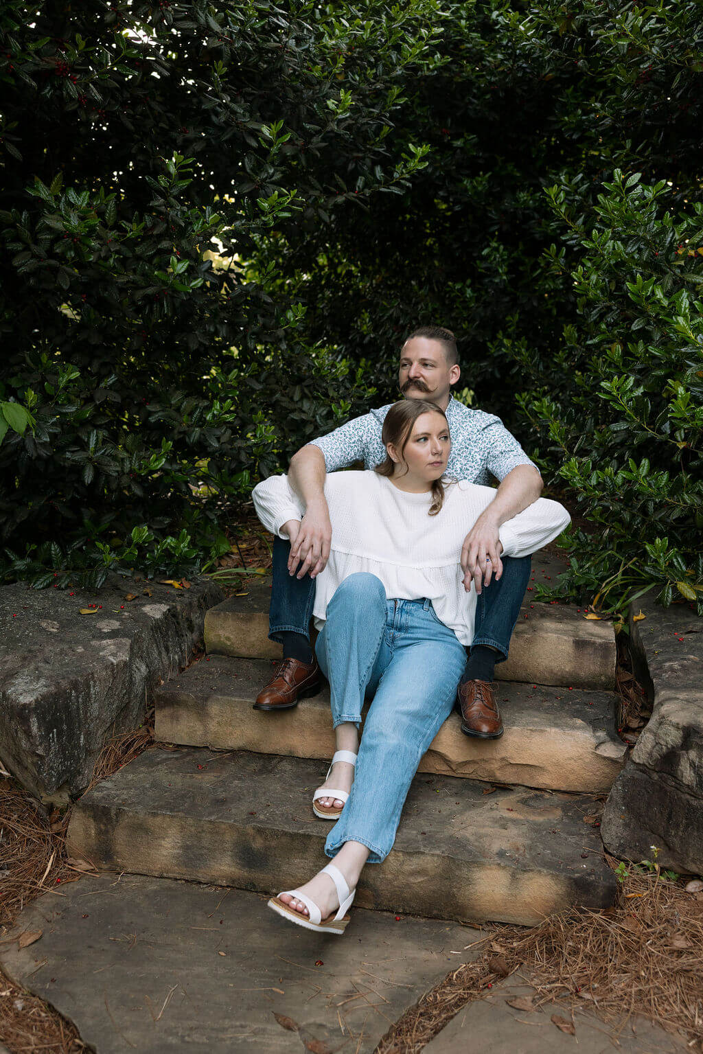 Engaged couple sitting together on stone garden steps at Knoxville Botanical Garden, relaxed and laughing, the woman in a white top and jeans leaning into her partner surrounded by lush greenery.