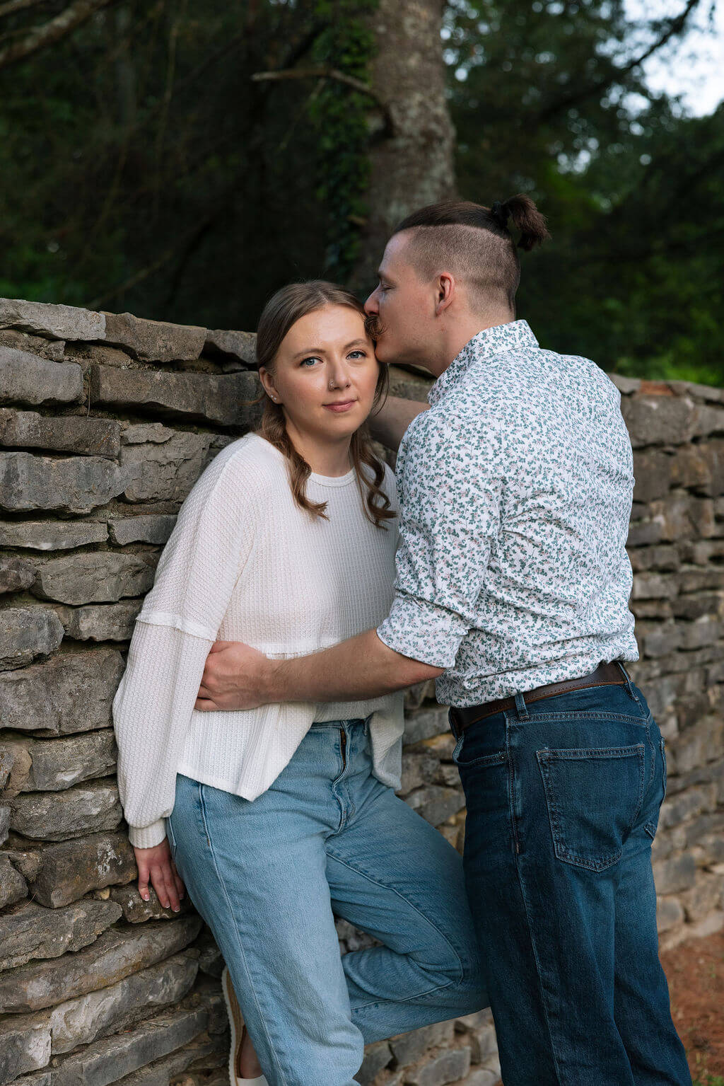 Couple leaning against a historic stone wall at Knoxville Botanical Garden, the man kissing his partner on the cheek as she gazes softly at the camera in the fading evening light.